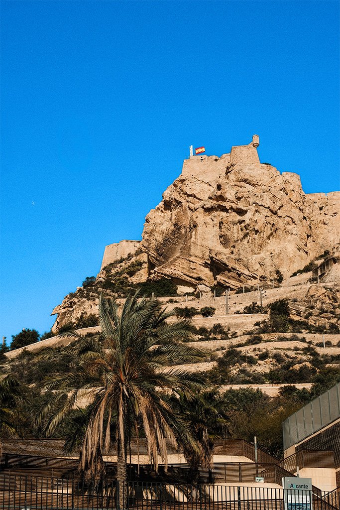 A view looking up at a rugged, tan-colored rocky mountain with a stone fortress perched at the summit, flying a Spanish flag. A large palm tree stands in the foreground, with zig-zagging pathways visible on the hillside below the castle against a bright, clear blue sky.